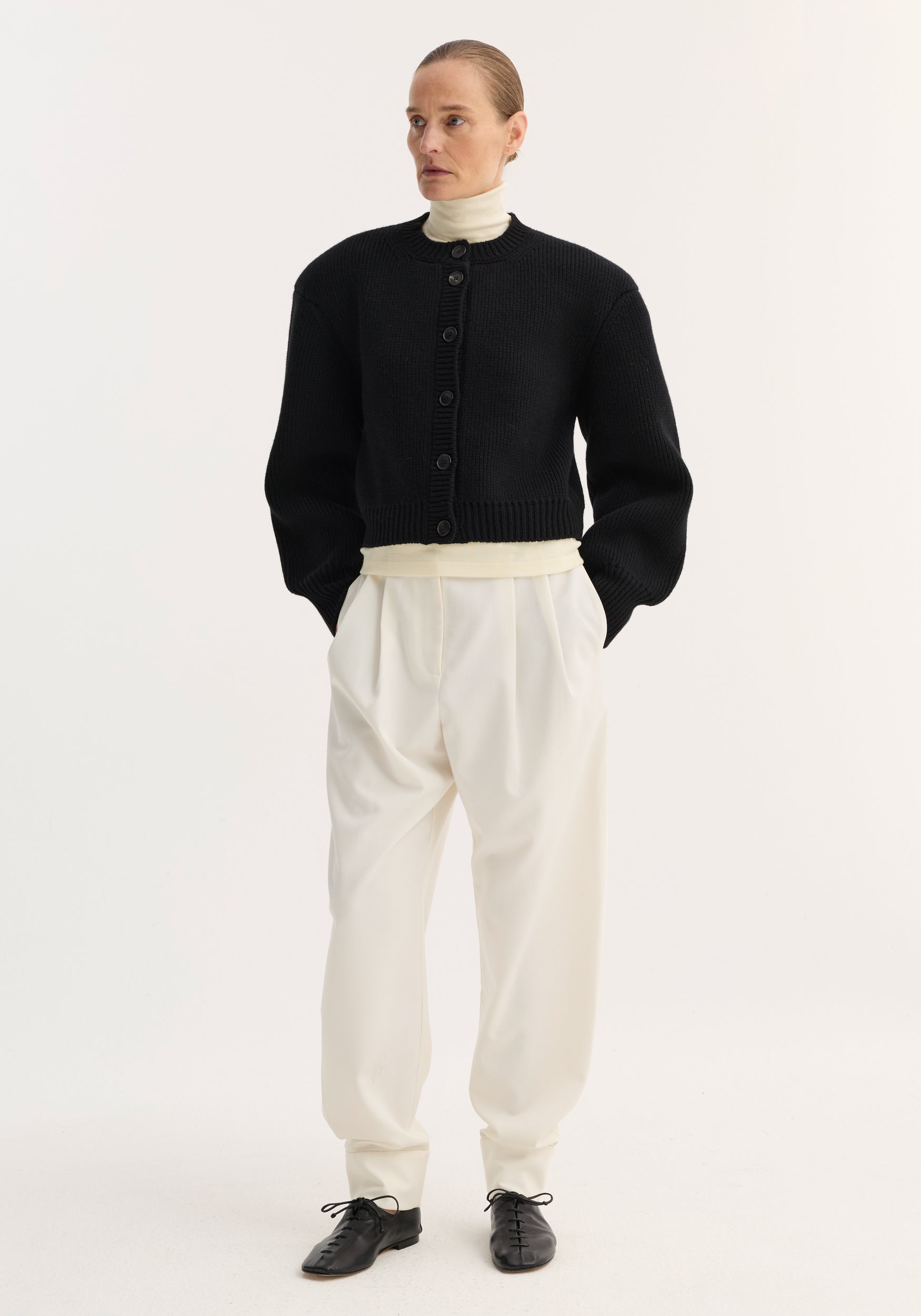Model wearing a black knitted Statement shoulder cardigan | black with oversized sleeves, paired with cream high-waisted pleated trousers and a cream turtleneck top.