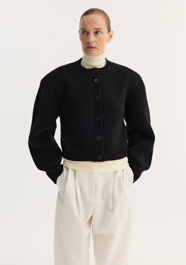 Model wearing a Statement shoulder cardigan in black, featuring an oversized fit and structured shoulders, layered over a cream top with wide-leg trousers.