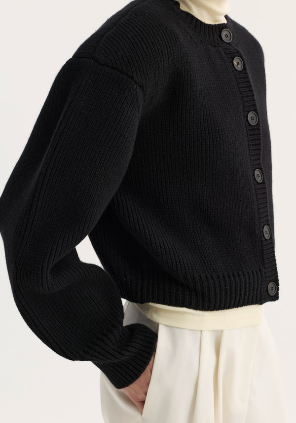 Statement shoulder cardigan in black, featuring ribbed-knit texture, ballooned sleeves, and button-front closure, layered over an ivory turtleneck.