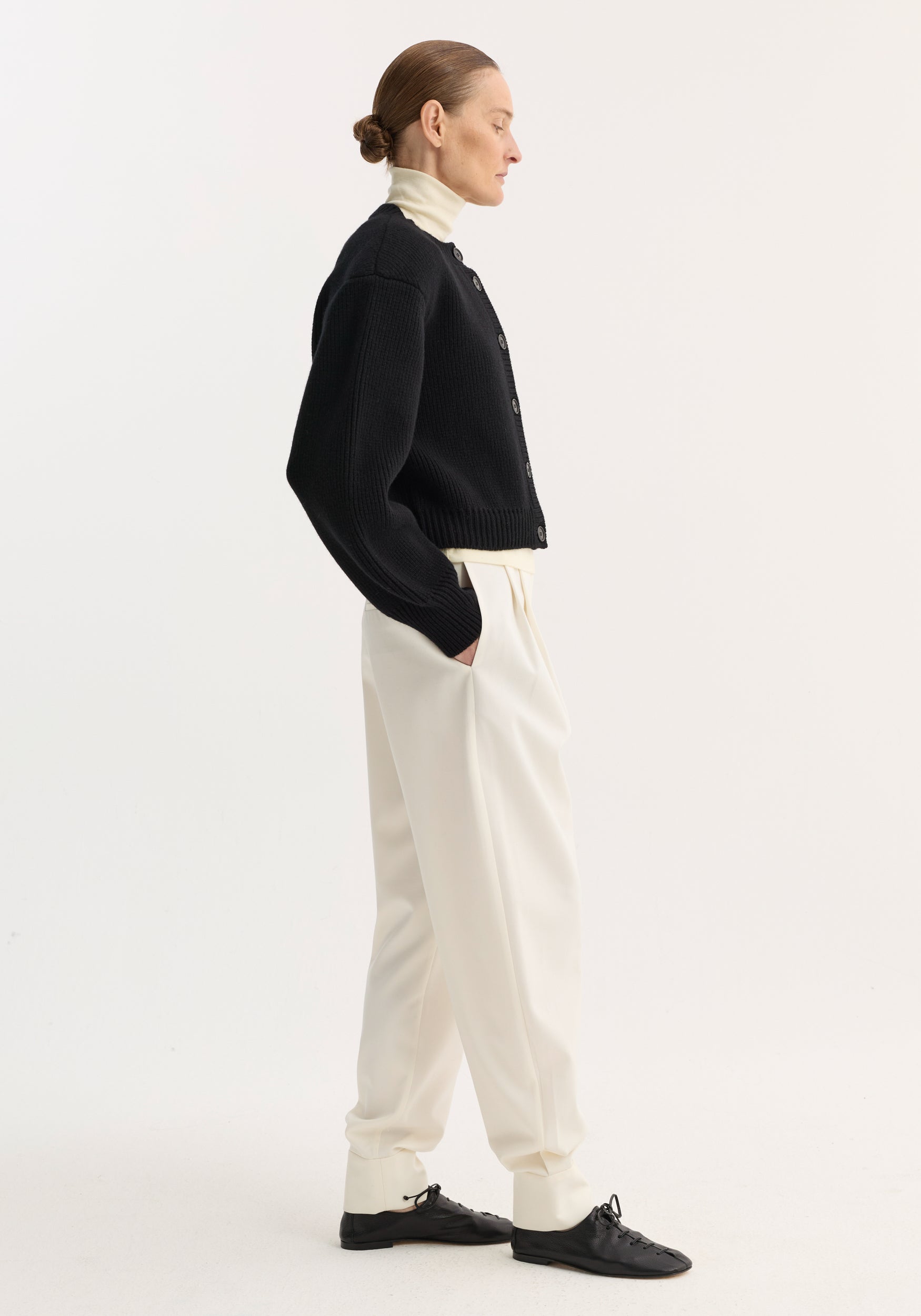 Side view of a woman wearing a black knit Statement shoulder cardigan | black over a white turtleneck, paired with white high-waisted pants and black lace-up shoes against a plain background.