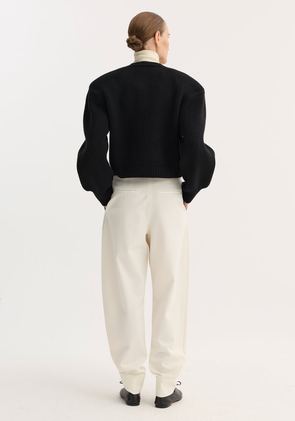 Back-view of model in black Statement shoulder cardigan with puffed sleeves, layered over a cream turtleneck, paired with high-waisted cream trousers and black flat shoes.