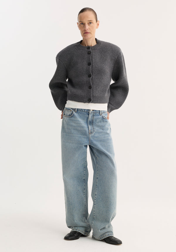 Woman wearing a mid grey melange Statement shoulder cardigan with dropped shoulders, over a white top and loose-fitting denim jeans, paired with black slip-on shoes.