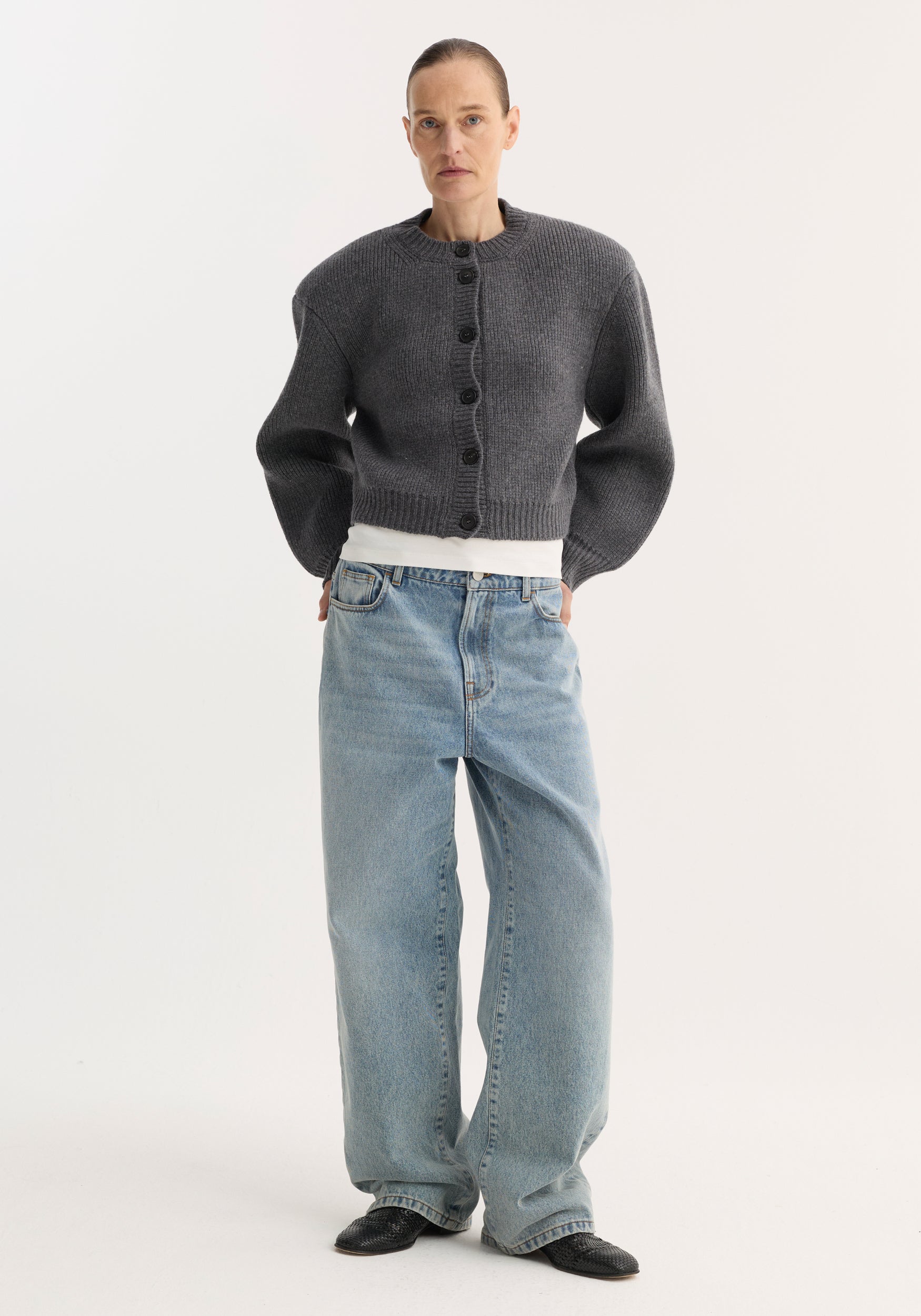 Woman wearing a mid grey melange knitted Statement shoulder cardigan with oversized sleeves and black buttons, paired with light blue wide-leg jeans and black shoes.