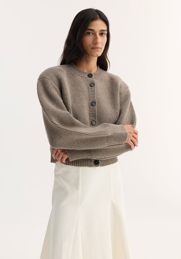 Young woman wears a Statement shoulder cardigan in clay melange, featuring a ribbed knit, button row closure, and oversized fit, paired with a high-waisted skirt.