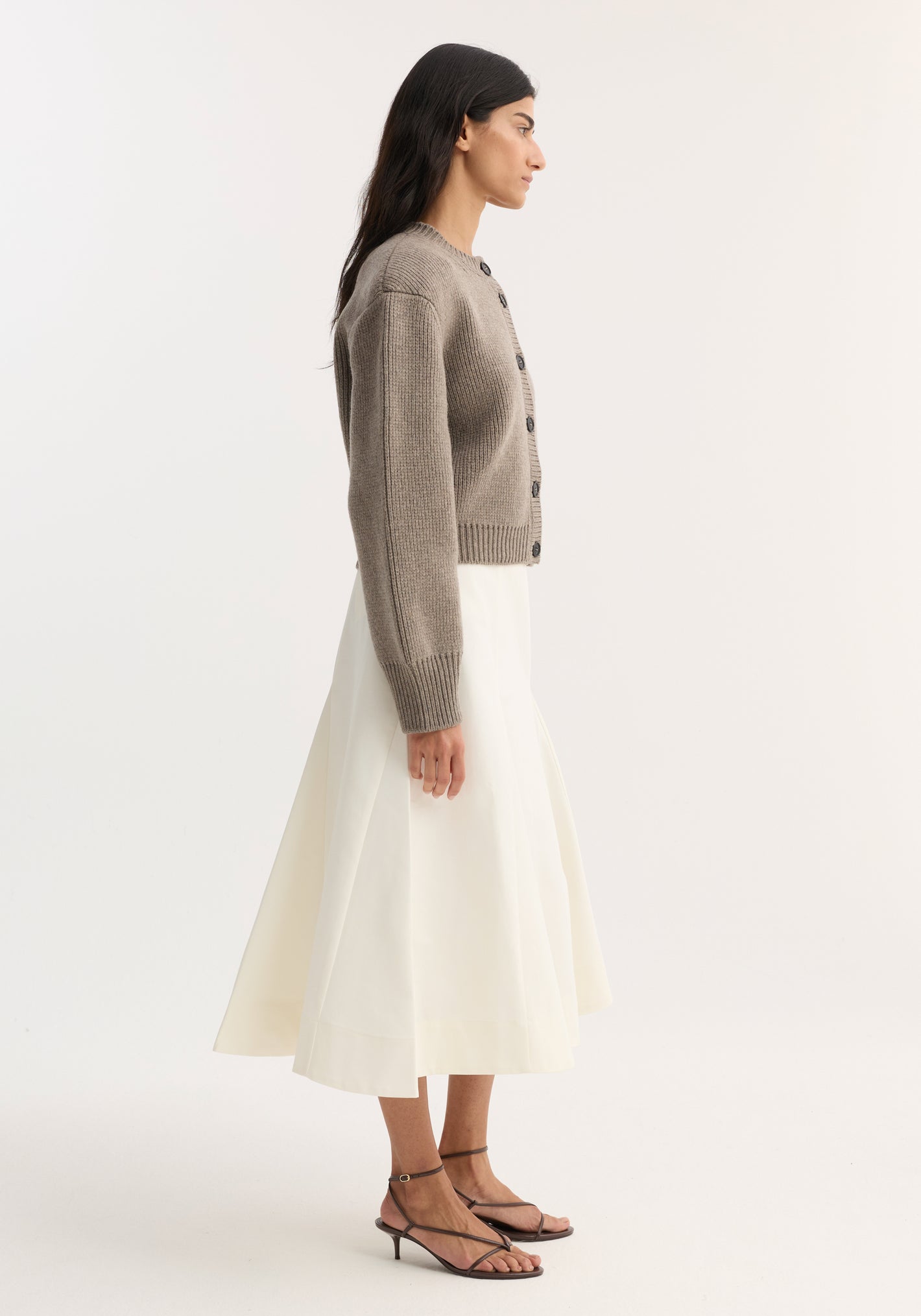 Woman in profile wearing a Statement shoulder cardigan in clay melange, featuring an oversized fit and button closure, paired with a midi skirt and strappy sandals.