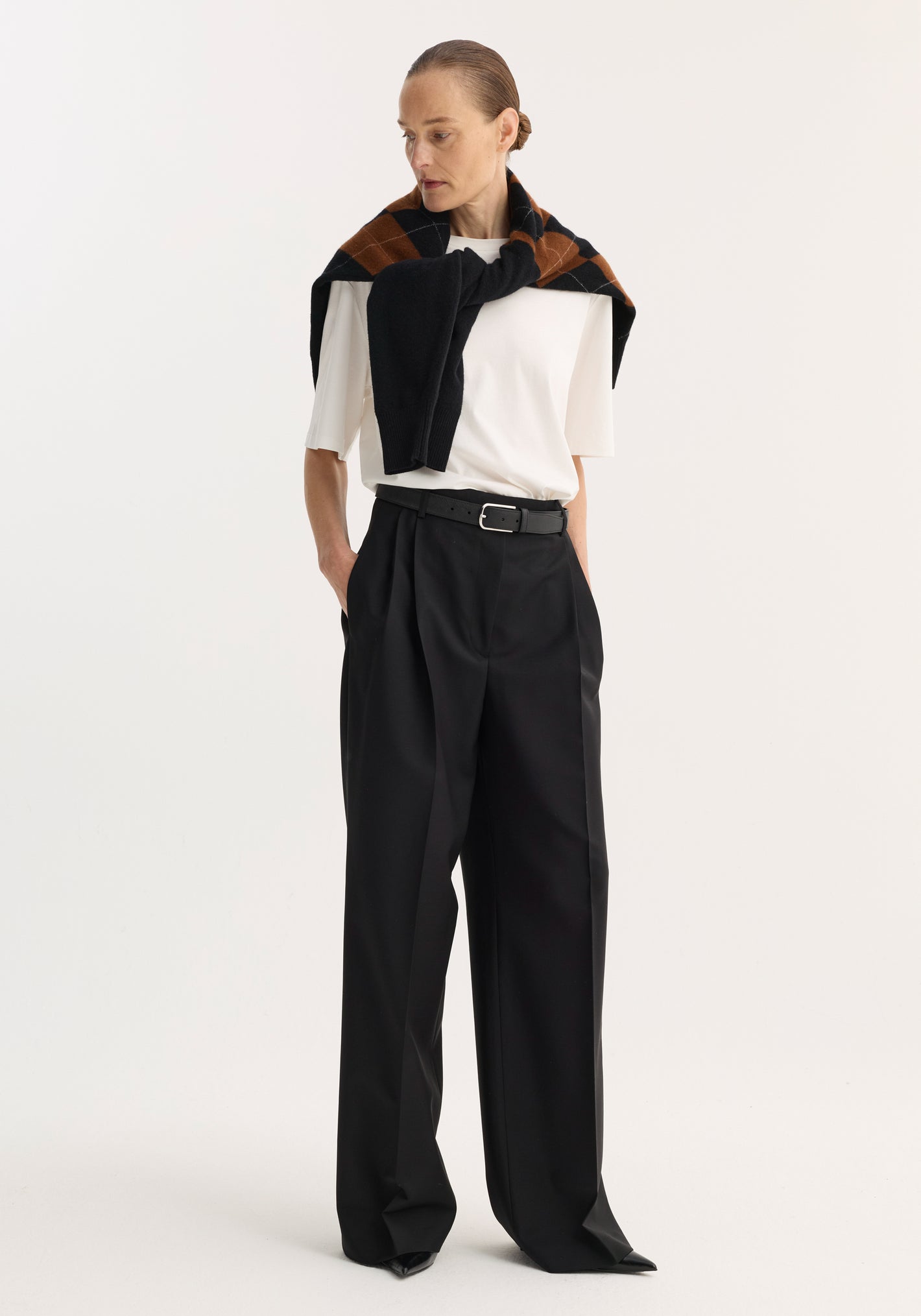 Woman wearing Mid-waist tailored trousers in black, featuring a wide leg and double pleats, complemented by a black belt and draped argyle-patterned sweater.