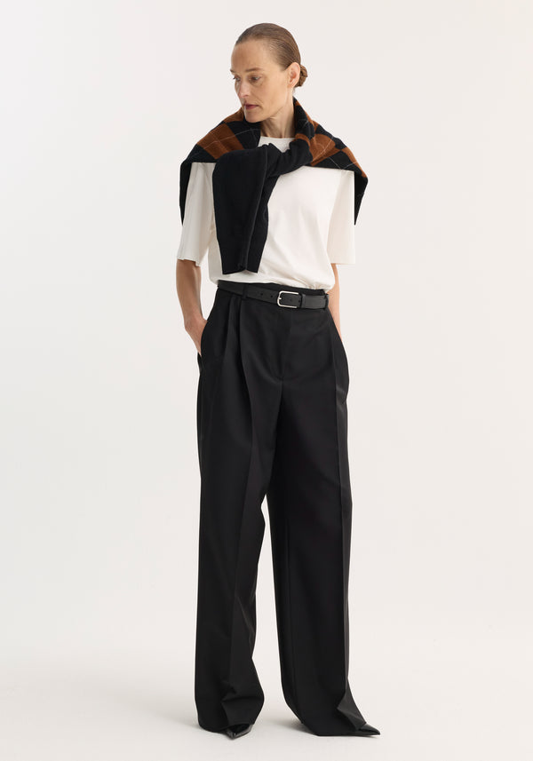 Woman wearing Mid-waist tailored trousers in black, featuring a wide leg and double pleats, complemented by a black belt and draped argyle-patterned sweater.