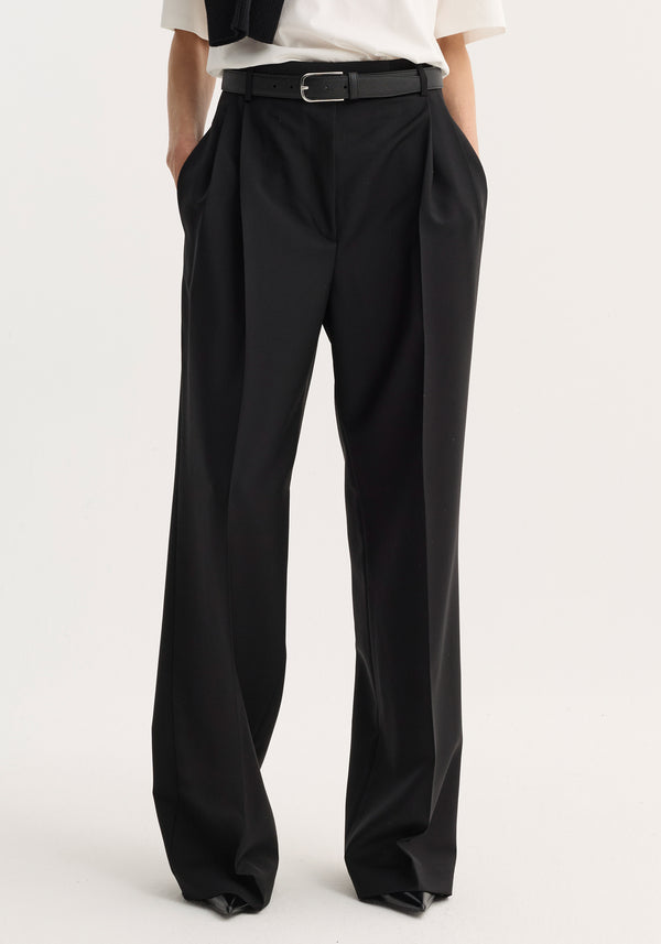 Mid-waist tailored trousers in black, featuring a high waist, wide legs, double pleats, and side pockets, worn with a belt and white tucked-in top.