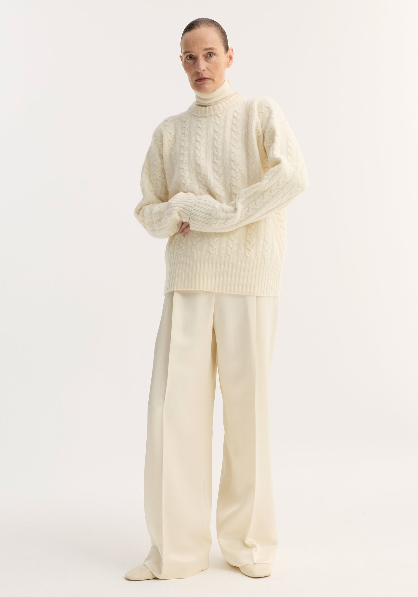 Person in off-white studio wearing Relaxed drawstring satin trousers in cream, featuring wide legs and drawstring waistband, paired with a chunky cable-knit sweater.