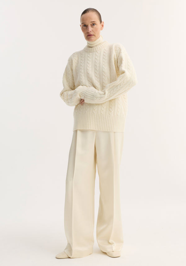 Person in off-white studio wearing Relaxed drawstring satin trousers in cream, featuring wide legs and drawstring waistband, paired with a chunky cable-knit sweater.