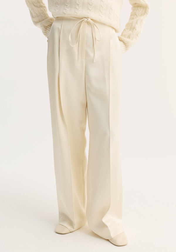 Relaxed drawstring satin trousers in cream, featuring a high-waisted, wide-leg design with pleats and a drawstring waistband, styled with ivory shoes.