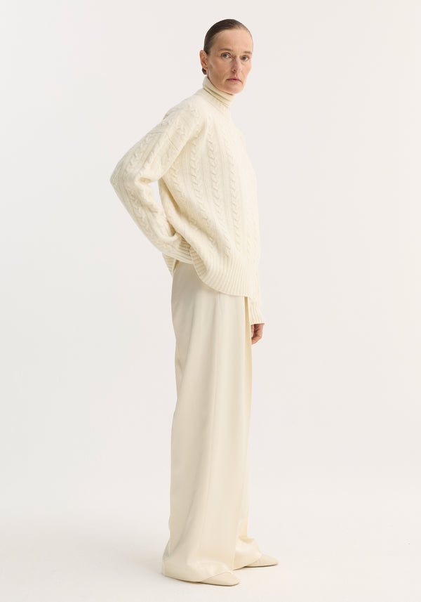 Woman in profile wearing cream relaxed drawstring satin trousers, featuring a long, wide-leg silhouette with front pleats, paired with an oversized cable-knit sweater.