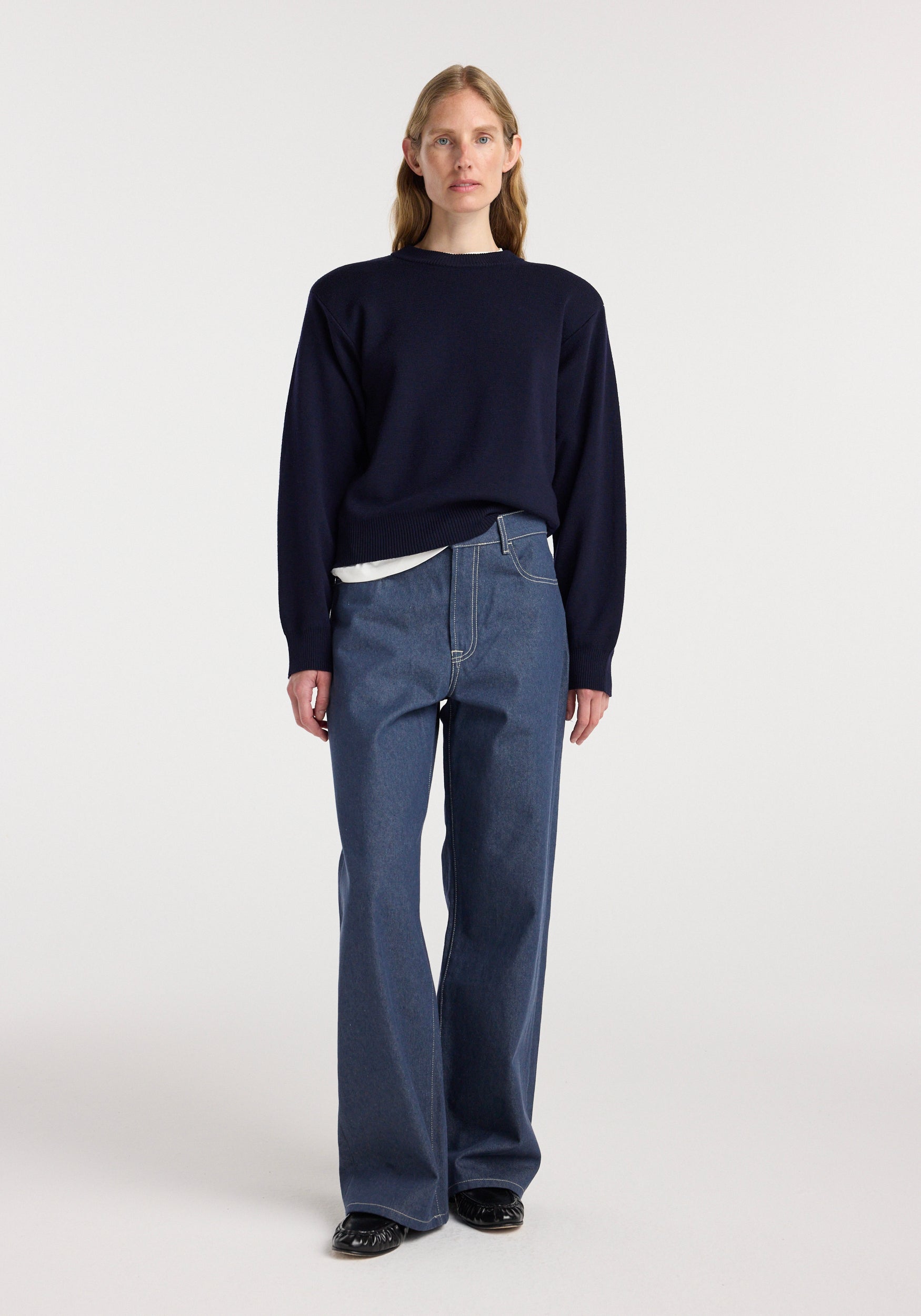 Woman wearing Bruun - Signature baggy denim | raw denim paired with a dark navy sweater and black shoes, standing against a plain light background.