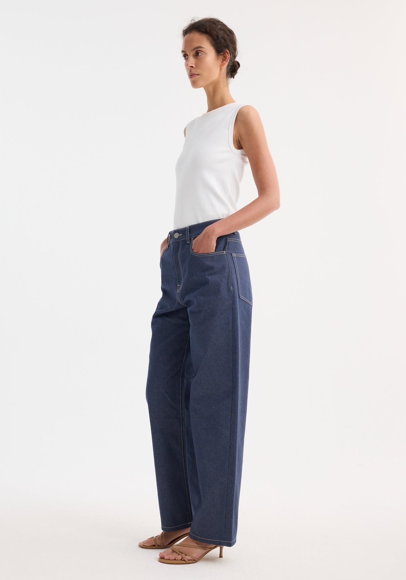 Model wears Signature baggy fit raw denim, showcasing a low-waist, wide-leg style. Paired with a white tank top and tan strappy sandals.