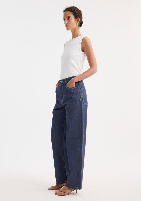 Model wears Signature baggy fit raw denim, showcasing a low-waist, wide-leg style. Paired with a white tank top and tan strappy sandals.