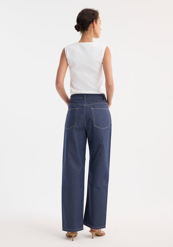 Model wearing Signature baggy fit raw denim, featuring wide-leg indigo jeans with contrast stitching, paired with a white rib-knit top and tan sandals.