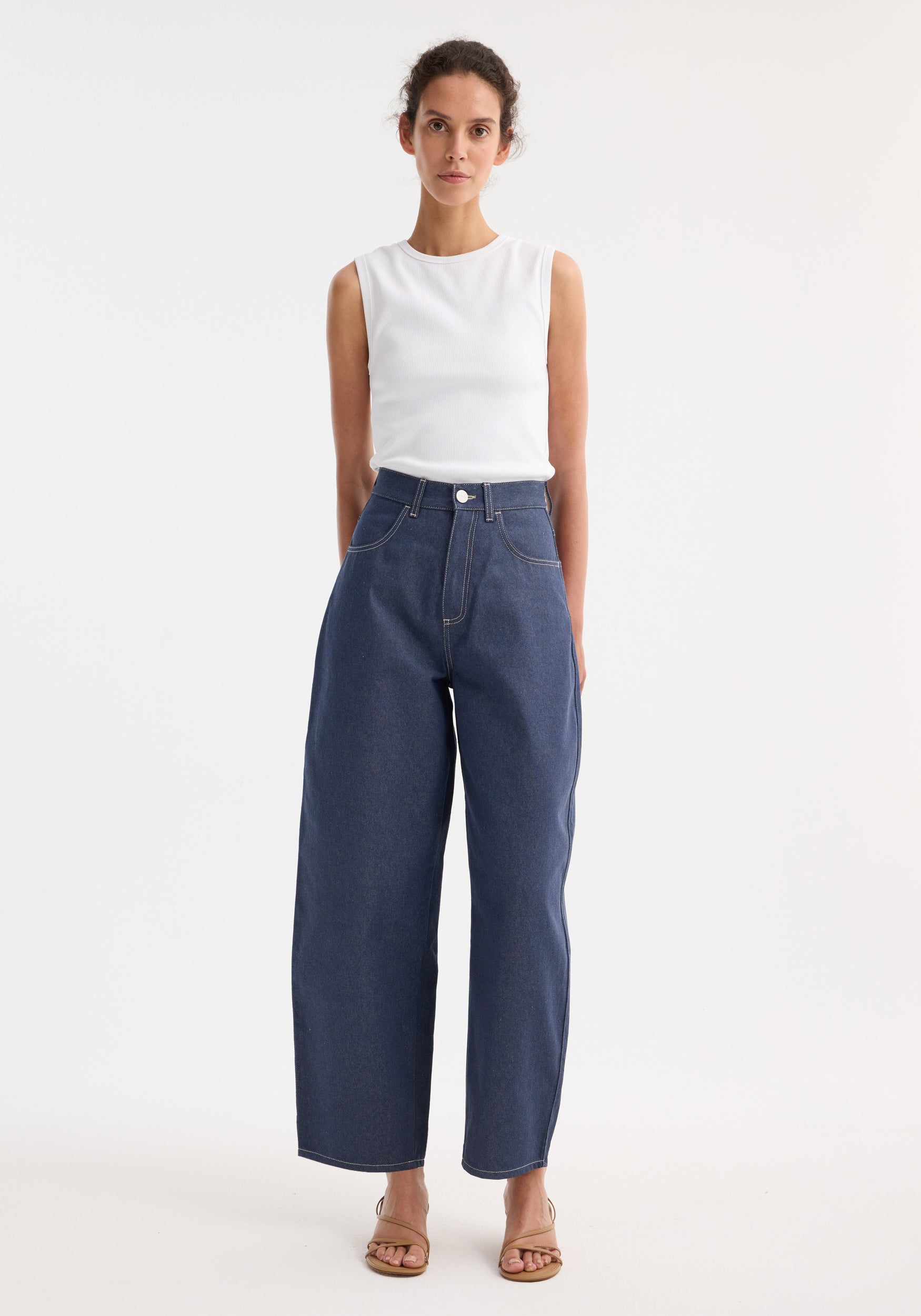 Model wears Barrel leg raw denim jeans with wide legs and heritage detailing, paired with a fitted white ribbed top and tan sandals.