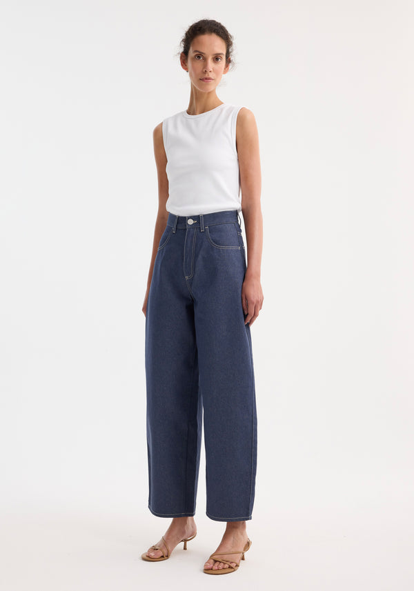 Model wearing Barrel leg raw denim with a white ribbed tank, showcasing wide-leg, high-waisted jeans in deep indigo, styled with tan strappy sandals.