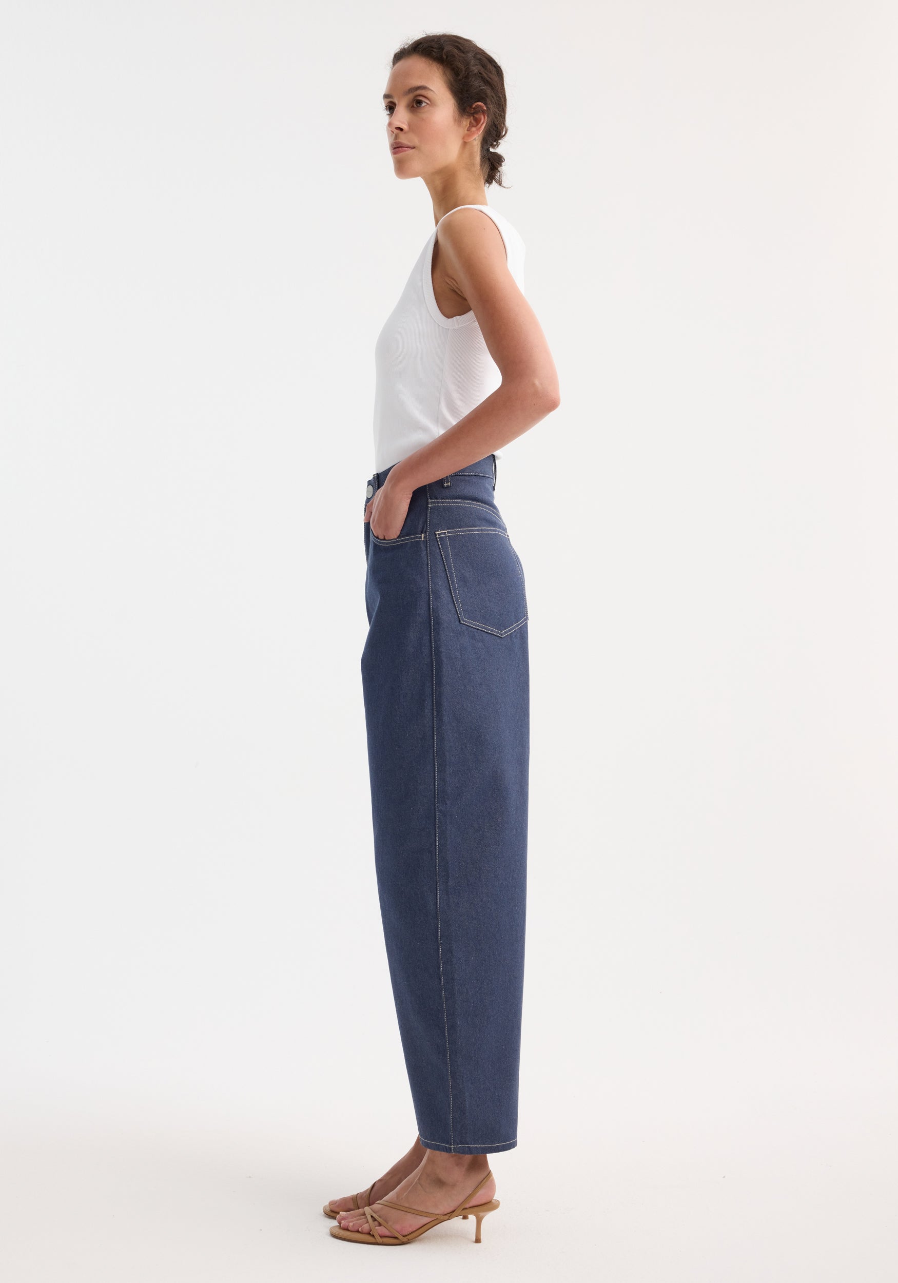 Barrel leg raw denim featuring a high-waisted, straight-leg design with heritage detailing, worn by a model in a white tank top and sandals.