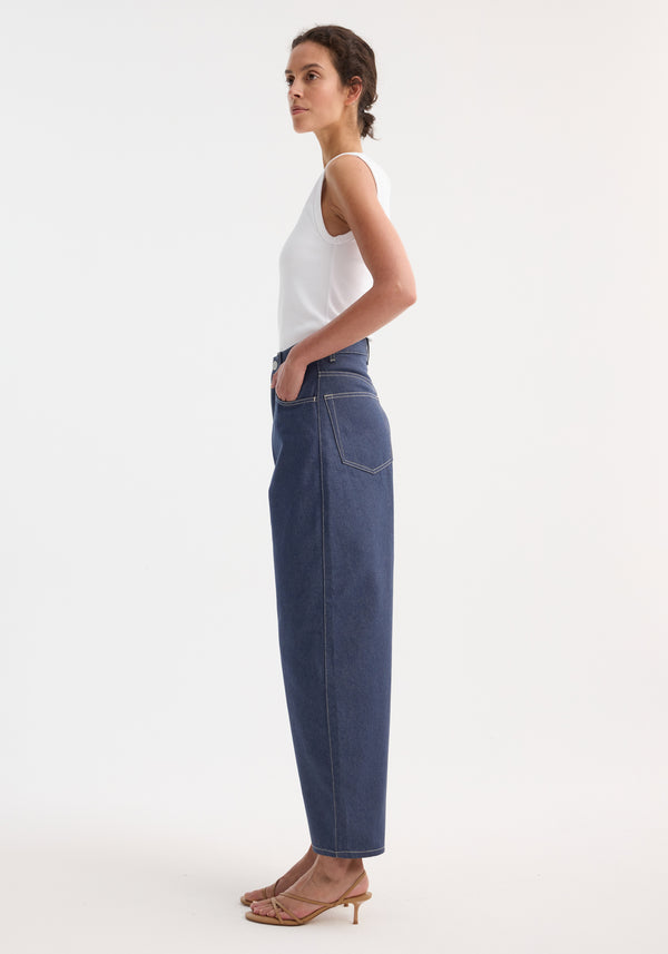 Barrel leg raw denim featuring a high-waisted, straight-leg design with heritage detailing, worn by a model in a white tank top and sandals.