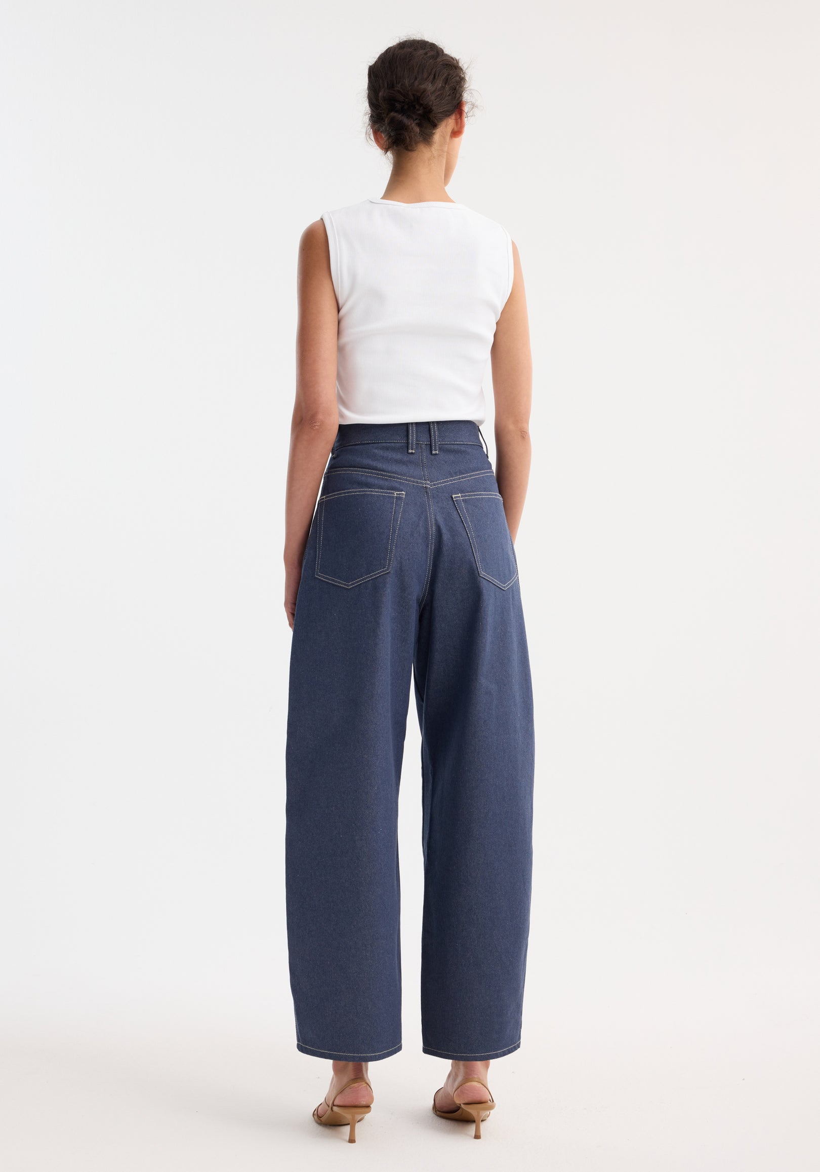 Person in a sleeveless top wearing barrel leg raw denim with wide legs, showcasing heritage detailing. Low-heeled sandals complete the modern, timeless look.