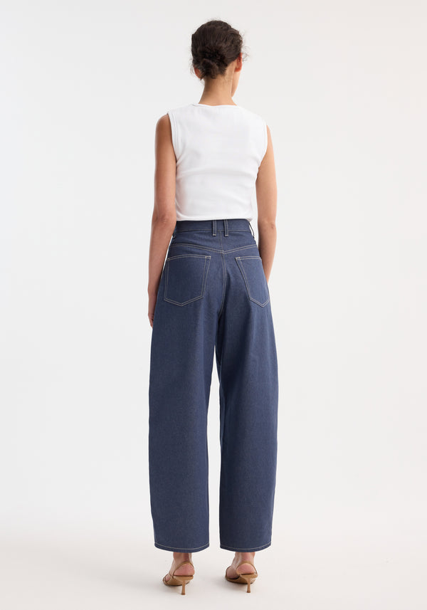 Person in a sleeveless top wearing barrel leg raw denim with wide legs, showcasing heritage detailing. Low-heeled sandals complete the modern, timeless look.