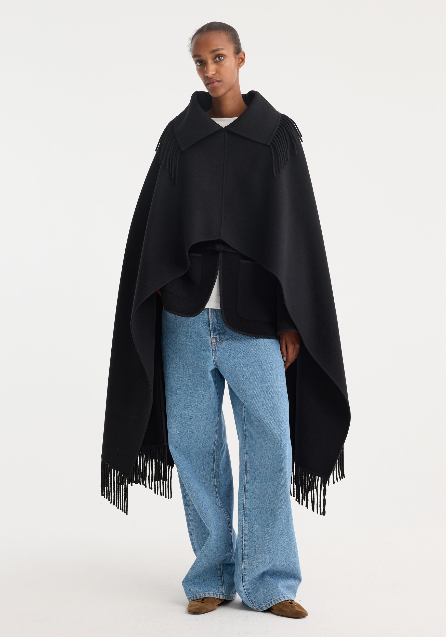Model wearing a Fringe poncho double-faced wool | black, featuring a wide collar, draping hem, and long fringed edges, over a simple white top and jeans.