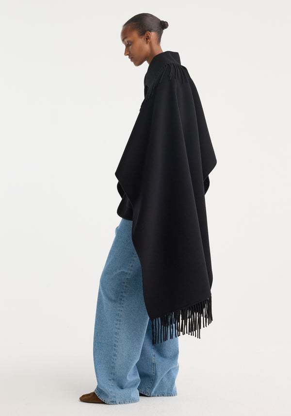 Fringe poncho double-faced wool in black, draped over a model with narrow fringe detailing, paired with light-blue jeans and brown-suede pointed-toe shoes.