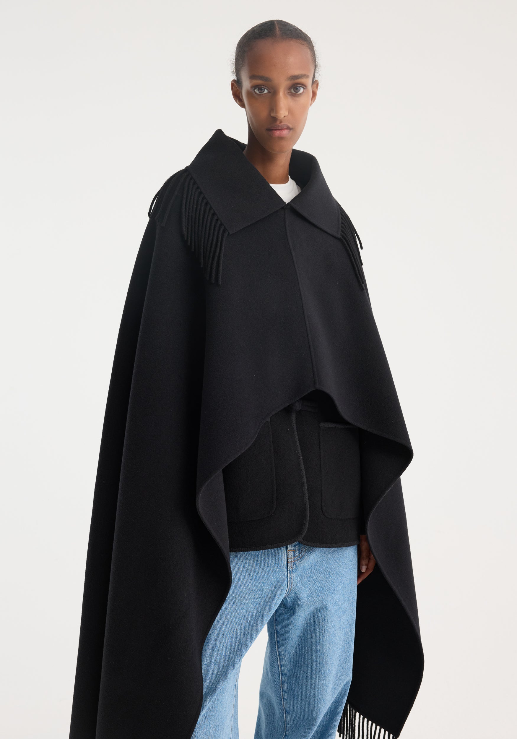 Fringe poncho double-faced wool | black, draped on a woman with fringed shoulders, over a black jacket and white top, paired with light-blue jeans.