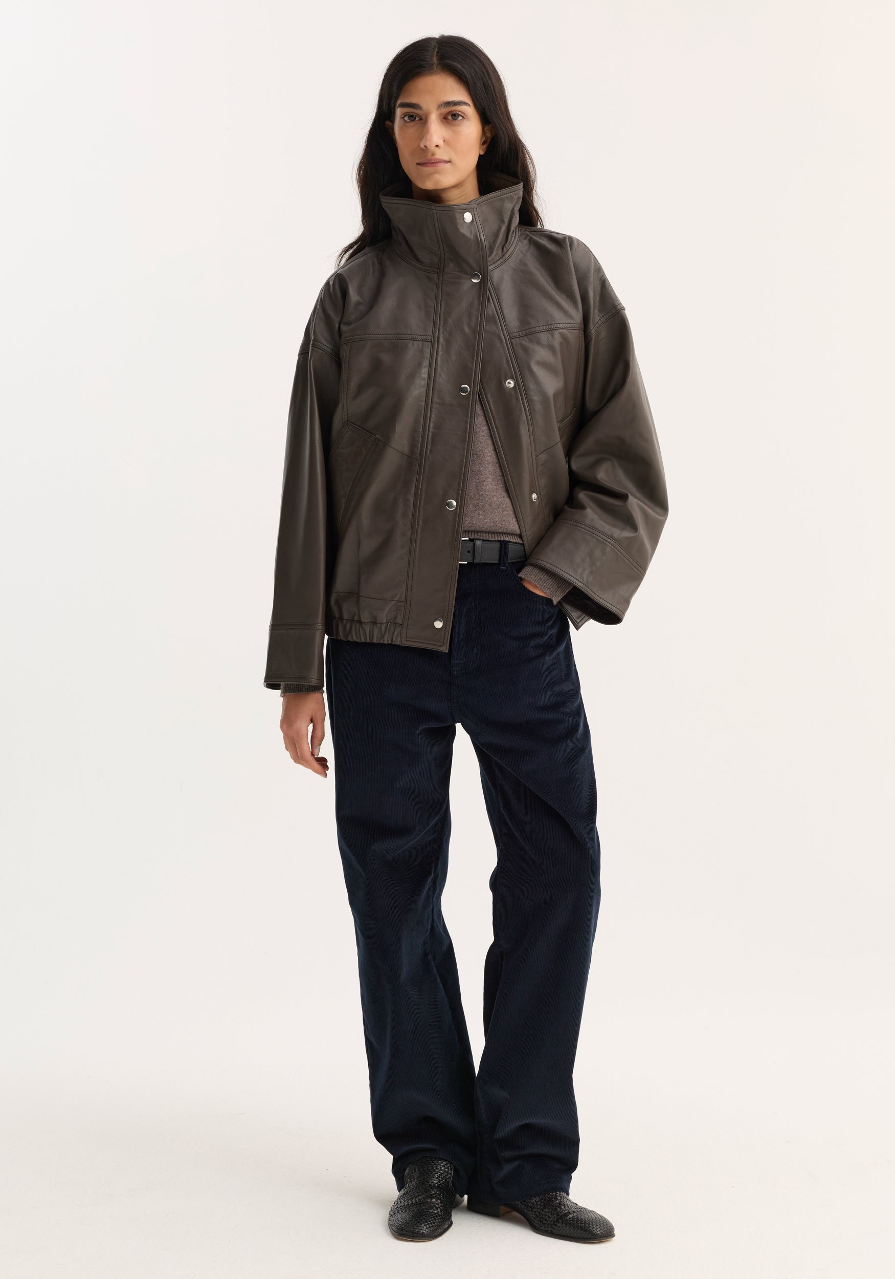 Woman wearing Reinvented leather jacket | earth, showcasing a boxy, dark-brown design with a stand-up collar, snap buttons, over navy wide-leg pants.