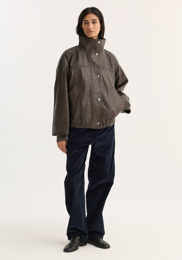 Woman wearing the Reinvented leather jacket | earth with a boxy, high-collar design and dropped shoulders, paired with navy wide-leg trousers and black low-heeled shoes.