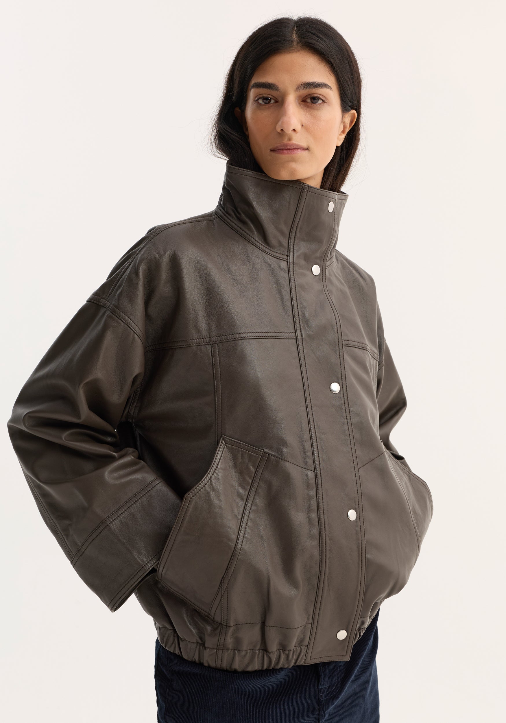 Woman wearing the Reinvented leather jacket | earth, showcasing its oversized fit, high stand collar, snap-button closures, and drop shoulders against a plain backdrop.