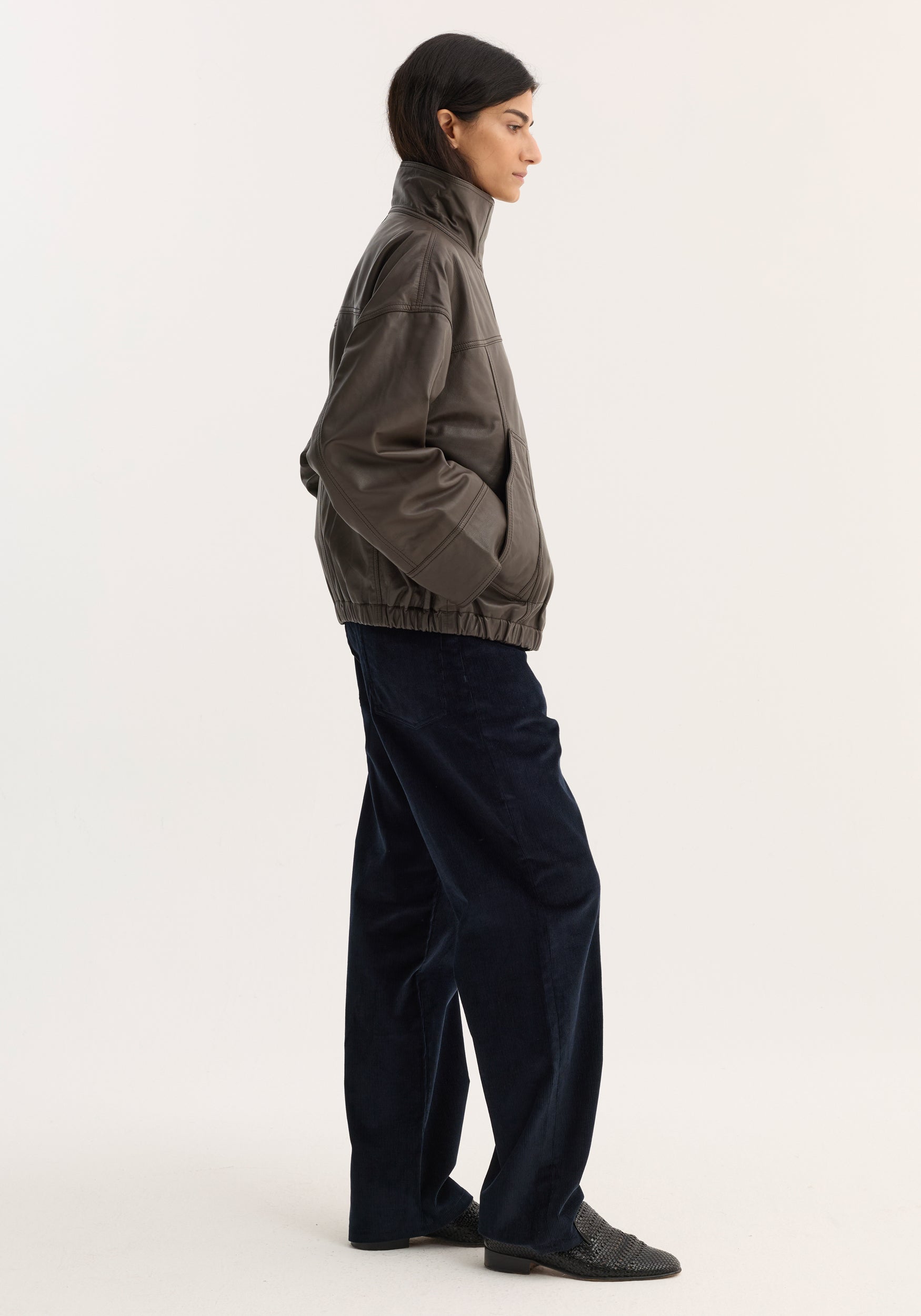 Reinvented leather jacket | earth: Profile view of a person wearing an oversized dark gray leather jacket with a high collar, paired with loose navy trousers and slip-on shoes.