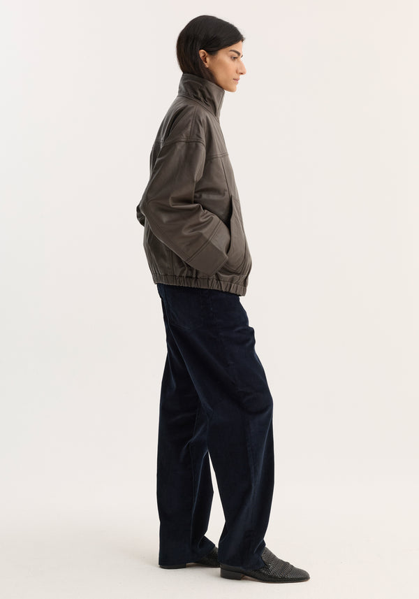 Reinvented leather jacket | earth: Profile view of a person wearing an oversized dark gray leather jacket with a high collar, paired with loose navy trousers and slip-on shoes.