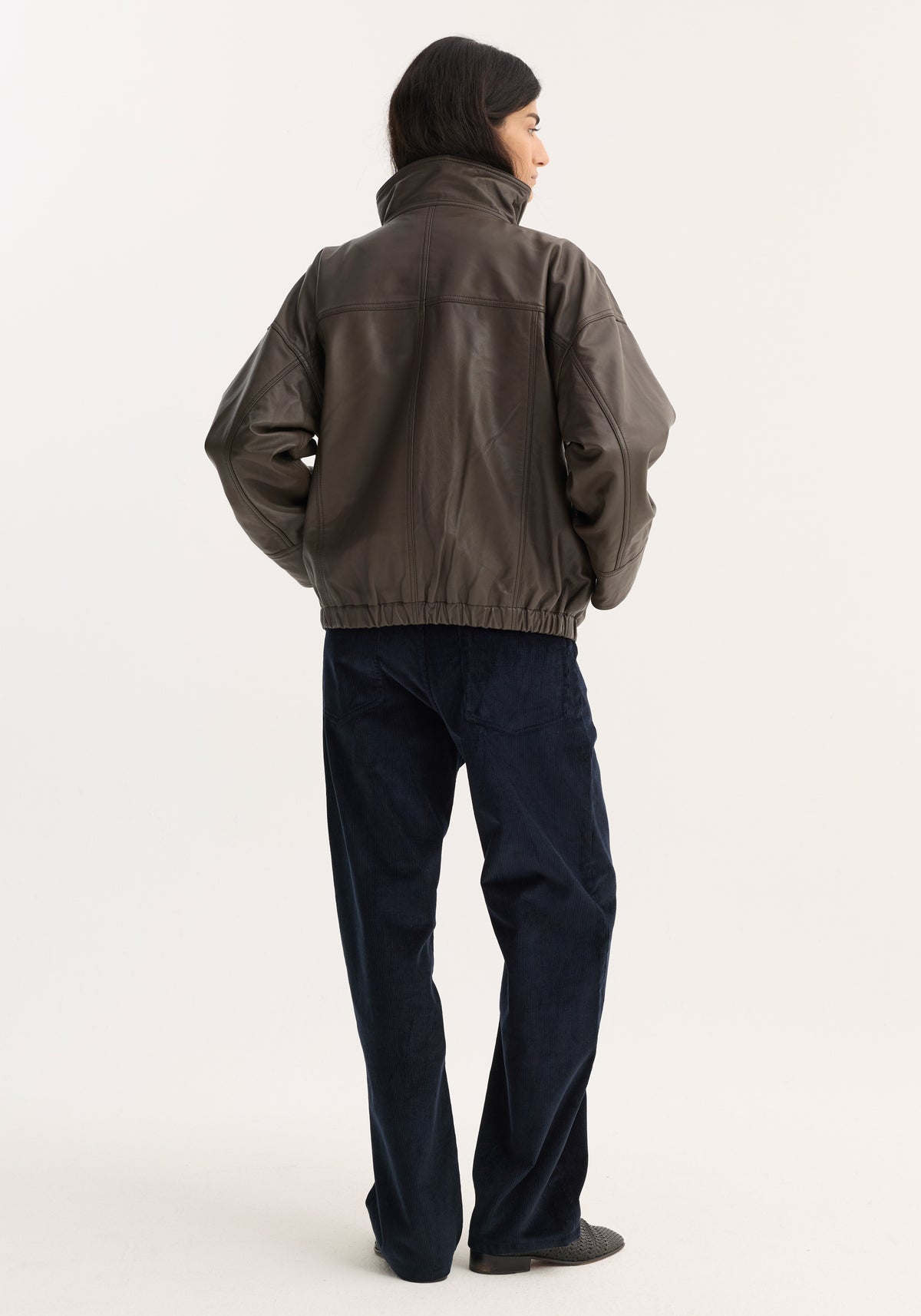 Person in an oversized Reinvented leather jacket | earth, featuring a high collar, elasticized hem, and snap closures, paired with navy trousers and slip-on shoes.