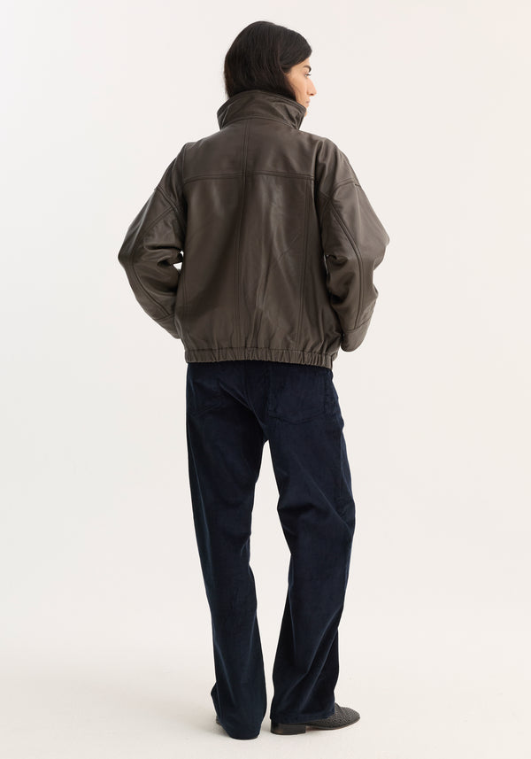 Person in an oversized Reinvented leather jacket | earth, featuring a high collar, elasticized hem, and snap closures, paired with navy trousers and slip-on shoes.