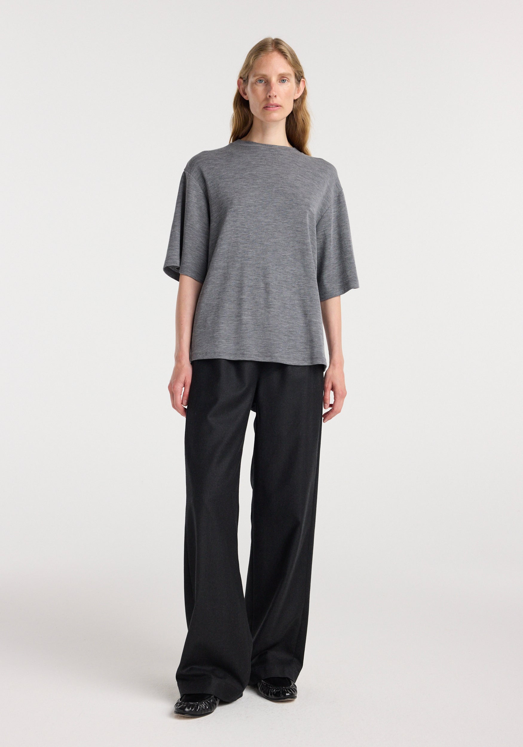 A woman wearing an oversized soft merino t-shirt | mid grey melange with short sleeves, paired with loose black trousers and black shoes, standing against a plain light background.