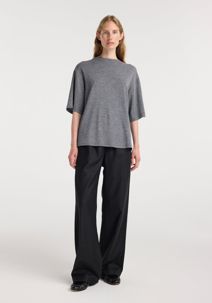 A woman wearing an oversized soft merino t-shirt | mid grey melange with short sleeves, paired with loose black trousers and black shoes, standing against a plain light background.