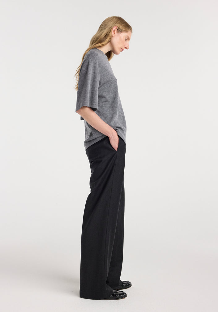 A woman wearing an oversized soft merino t-shirt | mid grey melange with short sleeves, paired with loose black trousers and black shoes, standing against a plain light background.