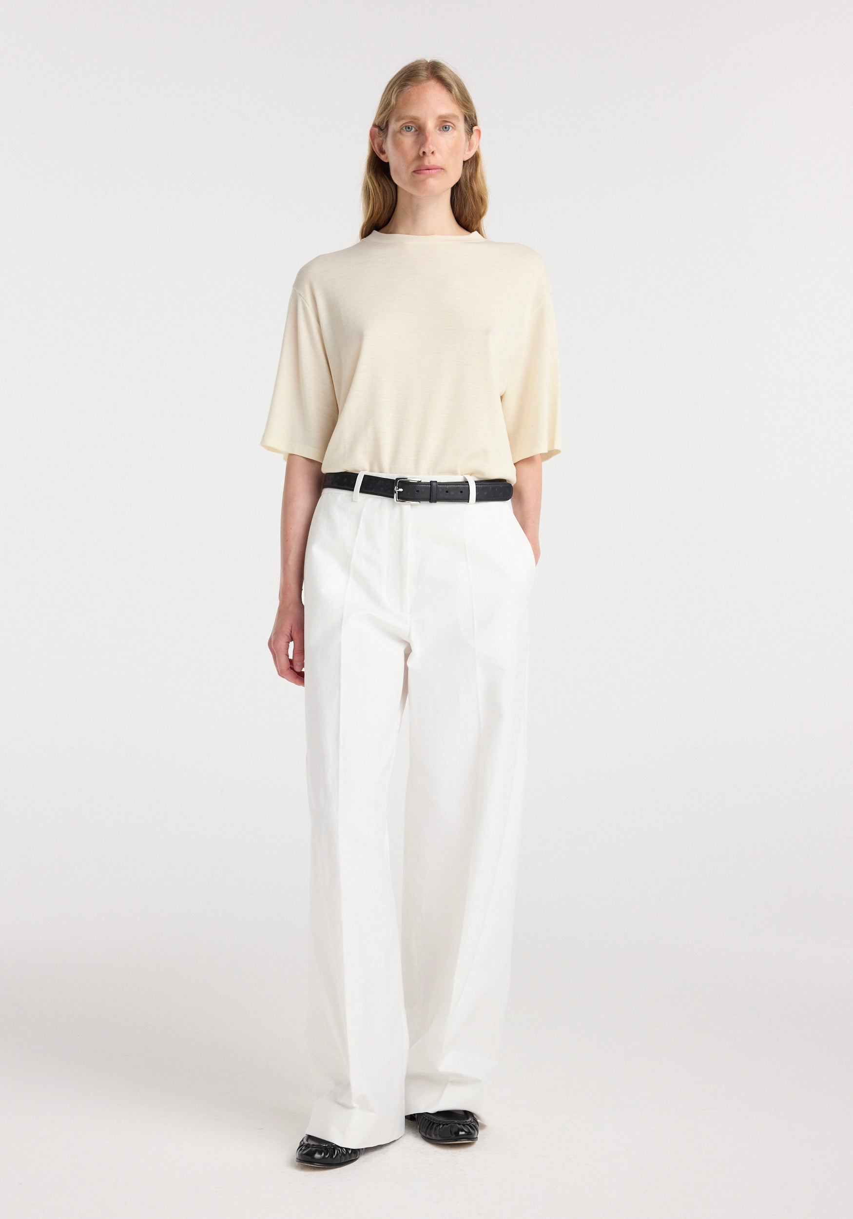 Woman wearing an oversized merino t-shirt | off white paired with white high-waisted trousers, a black belt, and black shoes, standing against a plain background.