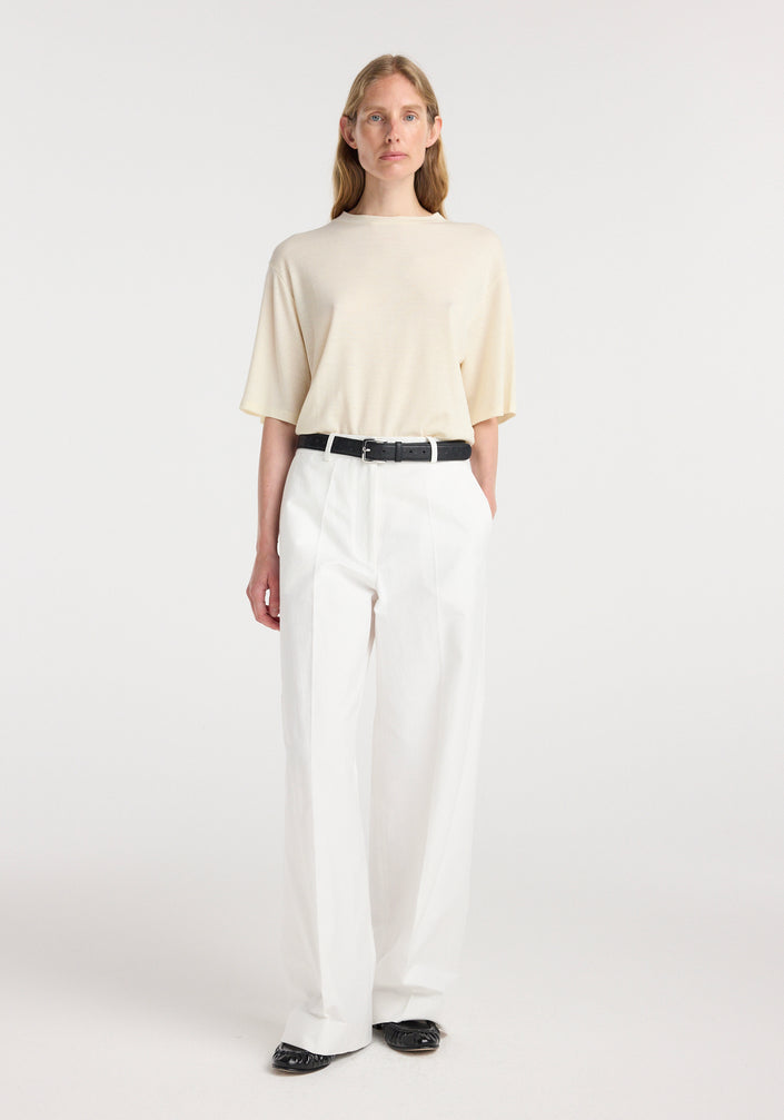 Woman wearing an oversized merino t-shirt | off white paired with white high-waisted trousers, a black belt, and black shoes, standing against a plain background.