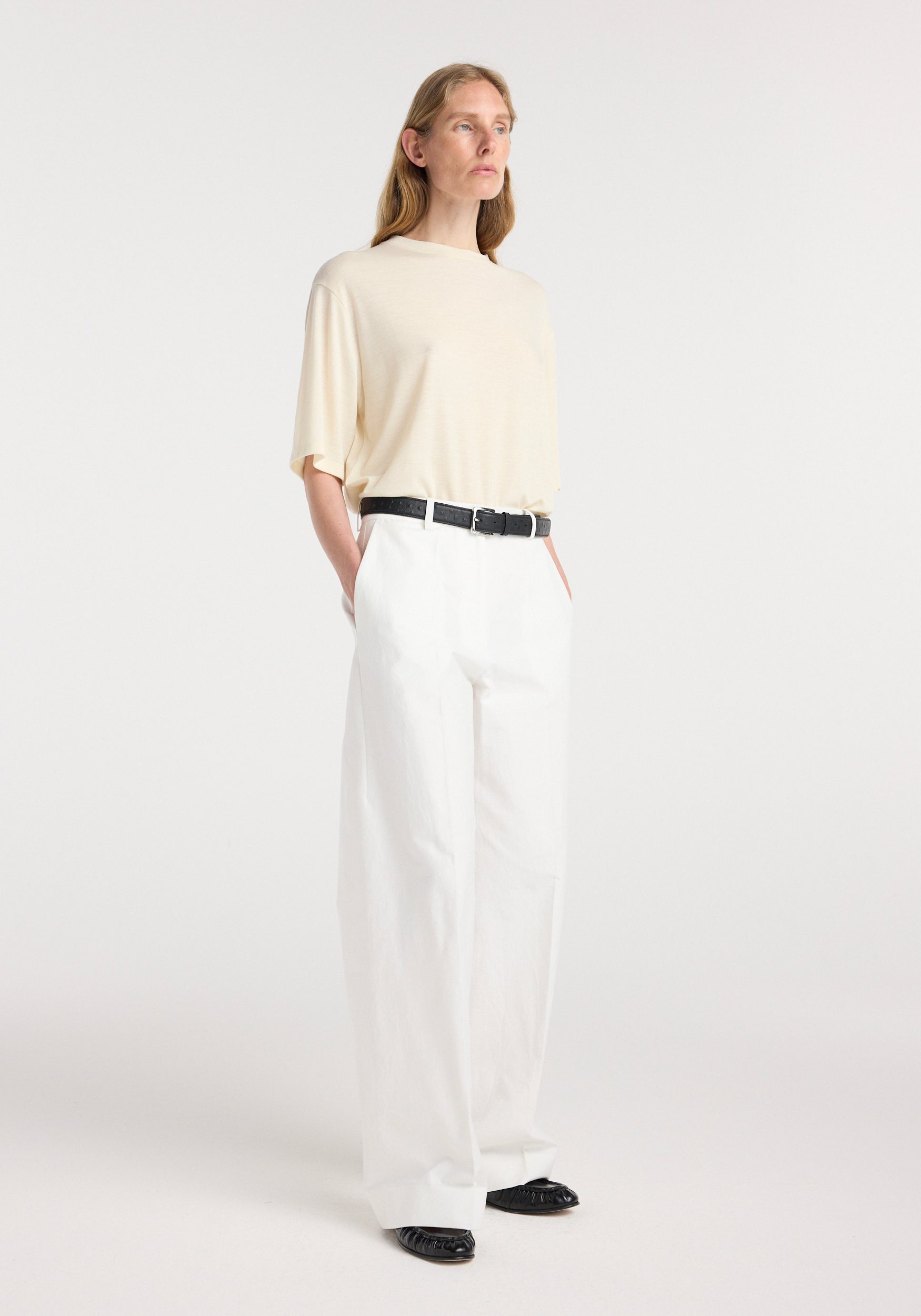 Woman wearing an oversized merino t-shirt | off white with wide-leg white pants and black belt, standing against a plain light background.