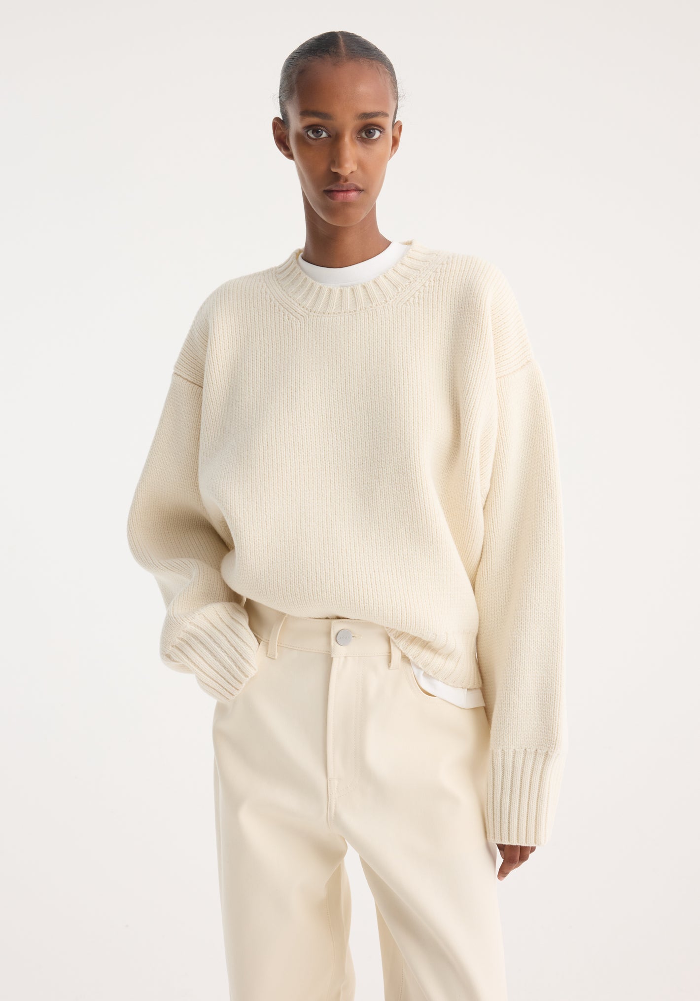 Person wearing a Sculptural compact crewneck in off white, showcasing a relaxed fit with dropped shoulders, paired with matching straight-leg trousers against a minimalist backdrop.