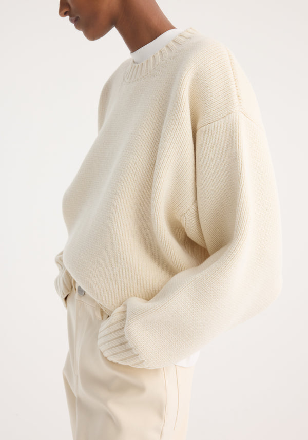 Side-view of a person wearing the Sculptural compact crewneck in off white, a loose-fit rib-knit wool sweater with dropped shoulders, paired with light beige trousers.