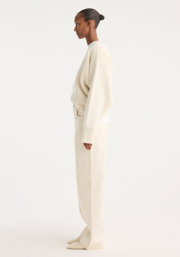 Model in profile wearing Sculptural compact crewneck, an oversized off-white knit sweater with drop shoulders, over wide-leg trousers, styled with pointed-toe flats.