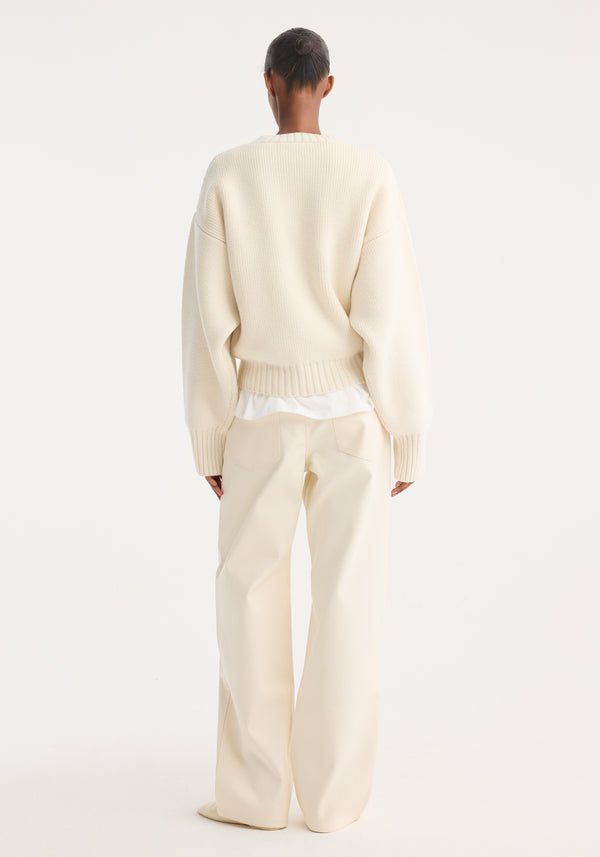 Person wearing Sculptural compact crewneck, an oversized cream knit sweater with dropped shoulders, over ivory trousers, hair in a bun, seen from behind.