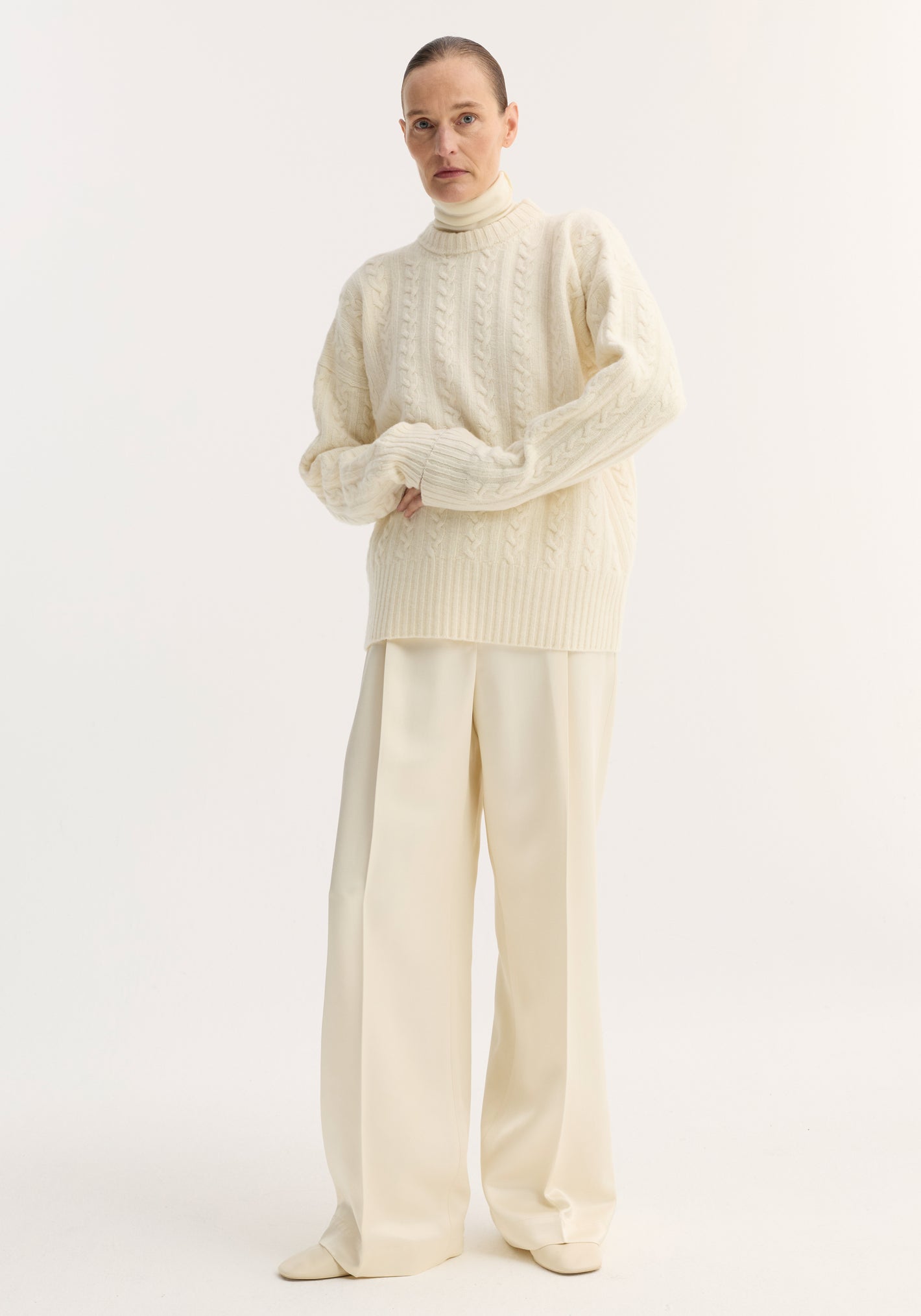Model in Cable knit jumper | off white: oversized, pure wool with dropped shoulders and round neckline, styled over a turtleneck and wide-leg trousers.
