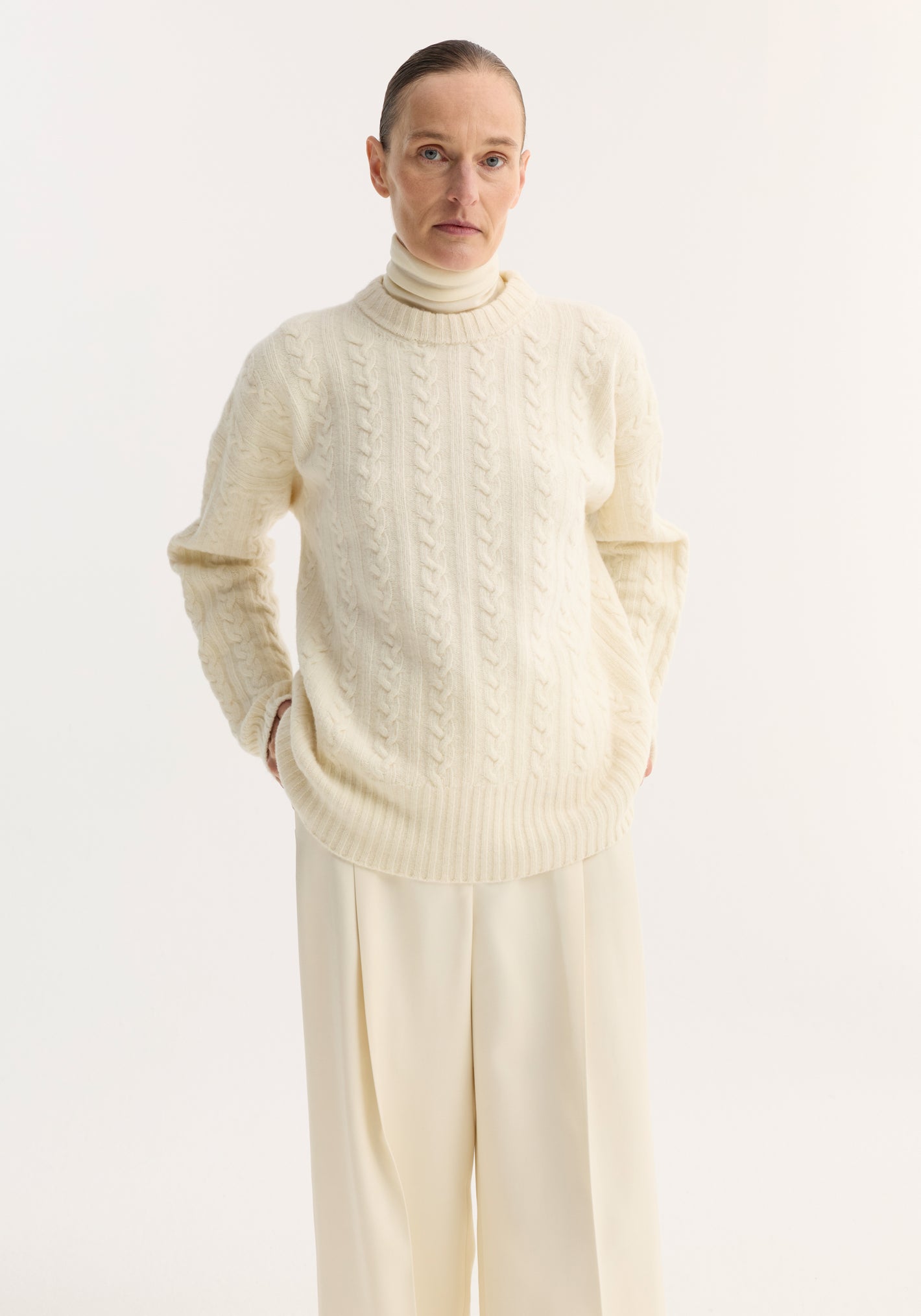 Person wearing an oversized Cable knit jumper | off white, featuring a classic cable pattern, round neckline, and drop shoulders, layered over a high-neck top.