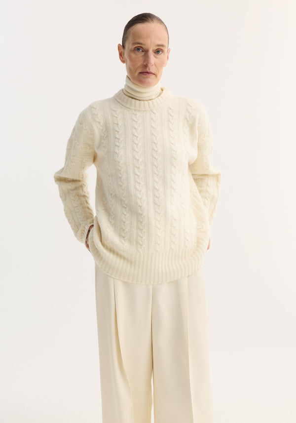 Person wearing an oversized Cable knit jumper | off white, featuring a classic cable pattern, round neckline, and drop shoulders, layered over a high-neck top.