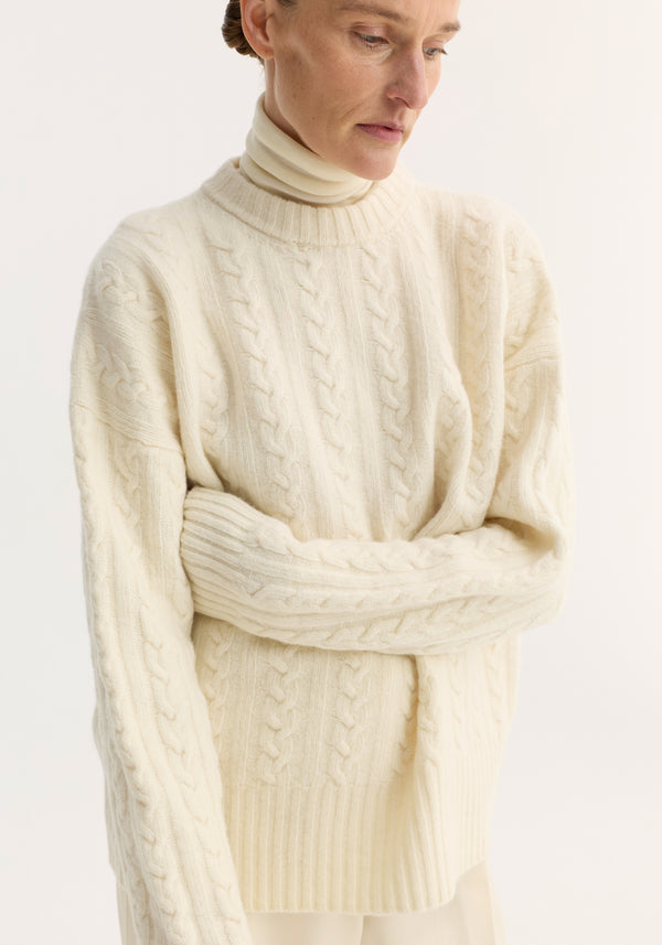 Cable knit jumper in off-white, worn by model with crossed arms. Features oversized fit, dropped shoulders, round neckline, crafted from pure wool for textured finish.